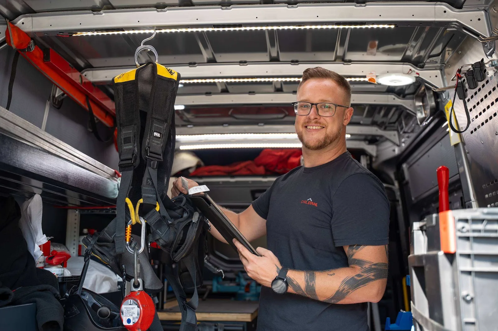 Servicetechniker im Carl-Stahl-T-Shirt mit Tablet im Servicefahrzeug, daneben ein Auffanggurt und ein Höhensicherungsgerät