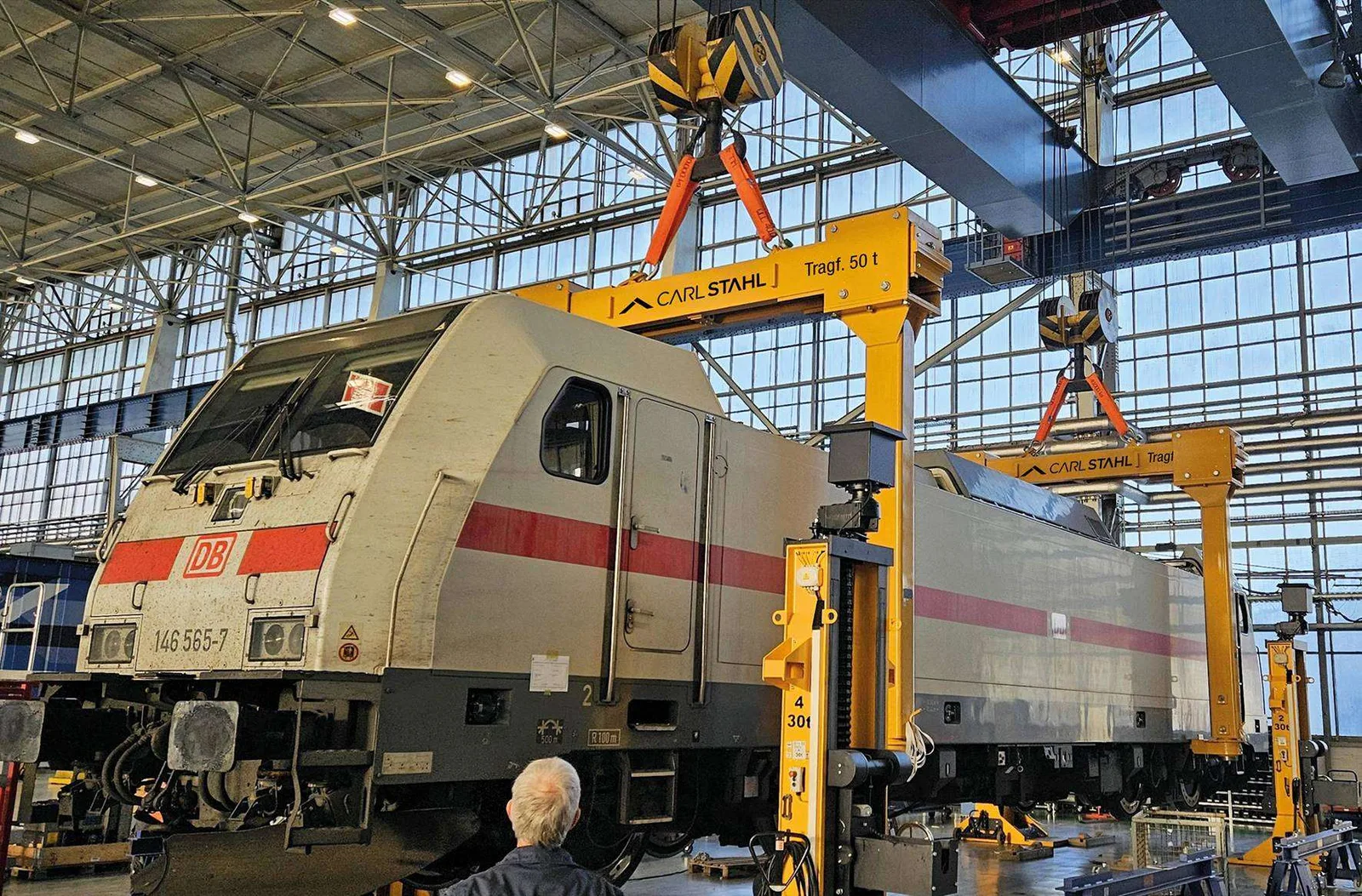 Eine Lok der DB hängt in einer Werkhalle an zwei gelben Carl-Stahl-Traversen mit der Aufschrift Tragf. 50 t