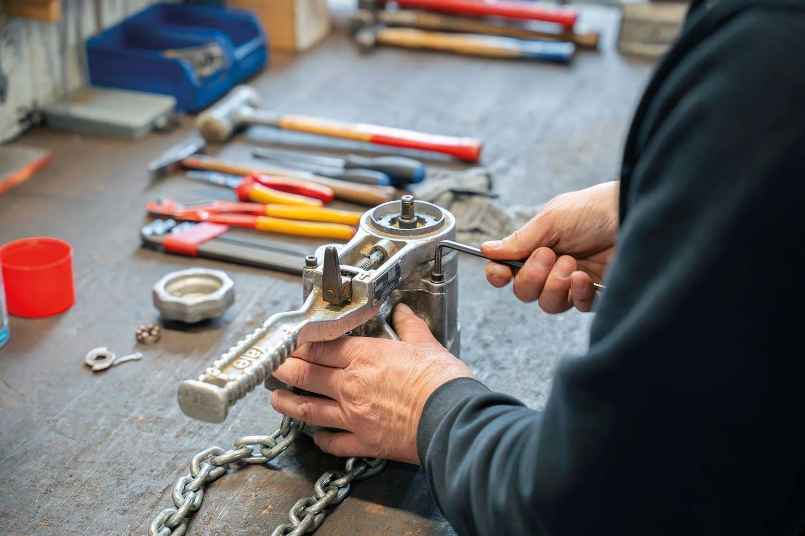 Hände reparieren einen Kettenzug an der Werkbank, daneben Hammer, Zangen und eine Kette